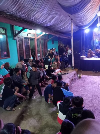 This nighttime image depicts a lively community gathering in Pangalengan, Indonesia, under a temporary tent structure with white and blue draped fabric. A large and diverse audience, consisting of men, women, and children of various ages, is seated informally on the ground and on concrete steps. Many women are wearing headscarves (hijabs).
The focal point of the event is a brightly lit, raised stage to the right, where a musical performance is taking place. Traditional Indonesian instruments, notably several kendang-like drums, are visible on the stage, alongside modern sound equipment such as speakers and amplifiers. A banner with partially discernible text, possibly starting with "PERINGATAN..." (meaning "Commemoration"), hangs in the background of the stage.
In the background, a house with a striking teal-colored exterior wall and traditional wooden-framed windows and doors is visible, suggesting the event is held in a residential or village setting. The ground where the audience is seated appears to be a mix of packed dirt and concrete. The atmosphere is engaged and communal, with artificial lighting illuminating the scene, creating a sense of celebration or shared entertainment.