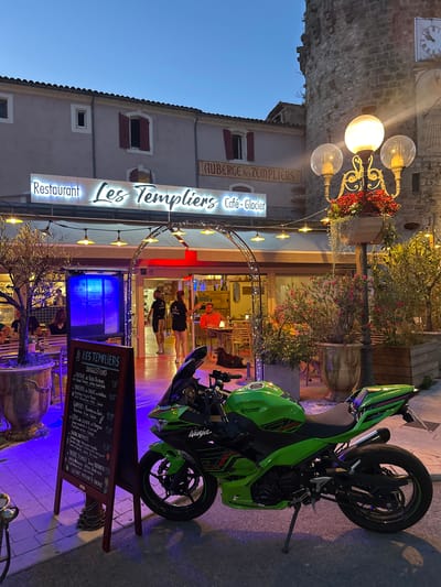 The image depicts an outdoor dining scene in front of a restaurant called "Les Templiers" in Anduze, France. The restaurant sign is illuminated with white lettering and is mounted above the entrance. Adjacent to the restaurant, a large stone tower stands, topped with a clock. The scene is set at dusk, with artificial lighting casting a purple hue on the ground and illuminating the restaurant's interior.

In the foreground, a bright green Kawasaki Ninja motorcycle is parked on the pavement. To the left of the motorcycle, a dark wooden A-frame sign displays menu suggestions for "Les Templiers," listing items like "Friture de Petits Poissons" and "Tartare de Boeuf au Gateau."

Several people are visible. Two individuals, a man and a woman, are standing near the restaurant entrance, appearing to be staff members. A few patrons are seated at tables outside the restaurant, enjoying their evening. A dog is also present, lying on the ground near the tables.

The environment includes potted plants and trees, as well as outdoor seating with tables and chairs. The overall atmosphere suggests a lively evening at a restaurant and bar.