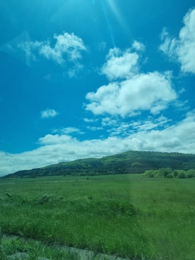 The image shows a view of the sky and ground, possibly taken from a moving vehicle. The sky is blue with scattered white clouds. The ground consists of a green field with a treeline along the edge. Based on the image location, this might be a view of the landscape in or near Relyovo, Bulgaria. There are no people visible in the image.