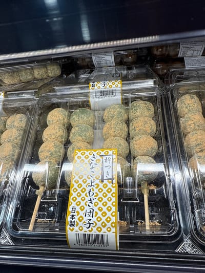 The image depicts a close-up view of several clear plastic containers stacked vertically, likely inside a refrigerated display case. The containers hold Japanese mochi balls, identified by a label with Japanese text and the words "よもぎ団子" (Yomogi Dango), which translates to mugwort dumpling. The mochi balls appear greenish and are coated with a tan-colored powder. Some containers feature mochi balls skewered on wooden sticks. A nutritional information label is visible on the top left corner. The barcode on the right shows a number of "4 982895 112197". The products are likely for sale in a store in Orlando, United States.