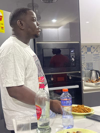 This vertical, eye-level indoor shot depicts a man in a modern kitchen, likely located in Adenta Municipality, Ghana. The subject is a Black man, seen in profile facing left, with short dark hair and a neatly trimmed beard. He is wearing a light-colored, short-sleeved t-shirt, and his gaze is directed slightly upwards.
His right arm is visible in the foreground, near two bottles. One is a clear plastic water bottle with a blue and orange label showing "PERLE." Next to it is a clear glass bottle with a red and black label, identifying the brand as "KASAPREKO" and the flavor as "RIVAL Cranyberry," with additional text "HERBA SAX." A clear plastic cup is positioned below these bottles.
The kitchen background features dark, built-in appliances, including an oven or microwave with a digital display clearly showing "10:00." Above the appliances, there is a white countertop and a decorative tiled backsplash with blue and white geometric patterns. A silver electric kettle is visible on the countertop further back. To the right, a yellow plate holds a serving of food, resembling fried plantains or a similar snack. A portion of a yellow and green energy efficiency label is visible on the far left, displaying "248" and the word "GHANA," which helps confirm the location. The scene suggests a casual moment in a domestic kitchen, possibly during meal preparation or a social gathering.