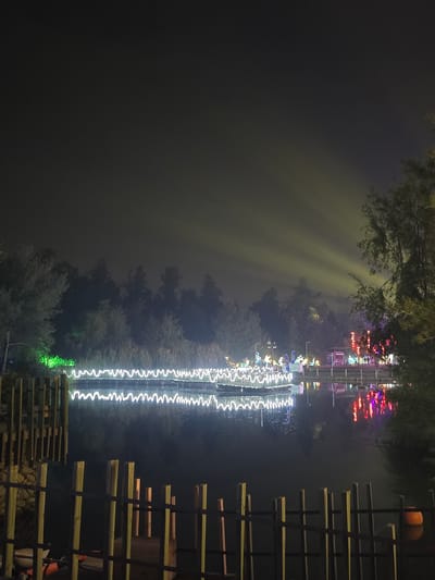The image shows a nighttime scene at Parque Pueblito de Las Vizcachas in Puente Alto, Chile. It features a body of water, possibly a lake or pond, with illuminated structures reflected in the water. These structures include a long, winding line of white lights resembling a bridge or pathway, and smaller structures with colorful lights.
A stack of wooden planks or pallets is visible in the foreground, along with a dark metal frame with horizontal wooden supports. At the top of the frame is a dark orange object, possibly a pumpkin or decorative gourd.
The surrounding environment includes trees and vegetation, with a hazy or foggy atmosphere obscuring details in the distance. The overall impression is of a festive or illuminated park display at night.