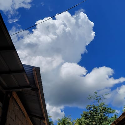 A wide-angle, low-angle shot captures a bright blue sky with dramatic cumulus clouds and a portion of a corrugated metal roofed structure in the foreground. The structure, appearing to be a building or shelter, is constructed with brick and wood, with a dark, weathered corrugated metal roof. A dark electrical wire stretches diagonally across the upper portion of the frame. In the lower right quadrant, the tops of lush green trees, including palm trees, are visible against the sky. The scene is set outdoors, likely in Samal, Philippines, given the tropical foliage and building materials. The lighting suggests it is daytime, with the sun illuminating the clouds and sky. There are no people visible in the image.