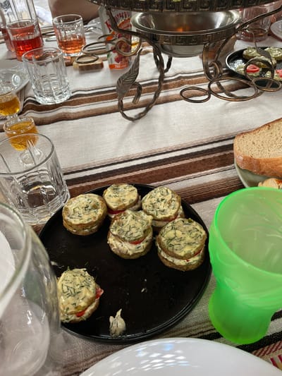 The image depicts a picnic scene with food and drinks laid out on a striped tablecloth. The subjects include several small, round sandwiches topped with a creamy sauce and herbs, a slice of bread, a green plastic cup, a black plate, and various glassware, some containing colored liquids. There is also a metal serving stand with decorative elements.

The activity appears to be a casual meal or gathering, possibly a picnic, given the setting and the array of food and drinks. The sandwiches, presented on a black plate, are the central food item. The glassware suggests beverages are being served.

Notable details include the close-up view of the food, highlighting the texture of the sandwiches and the dill in the sauce. The lighting suggests it might be daytime, though the time of day is not explicitly clear. The location is identified as Kobeliaky, Ukraine, which provides geographical context. No specific emotions are conveyed, but the scene suggests a relaxed, informal occasion.

Visible text includes "250g" on a small orange and white package visible on the serving stand.