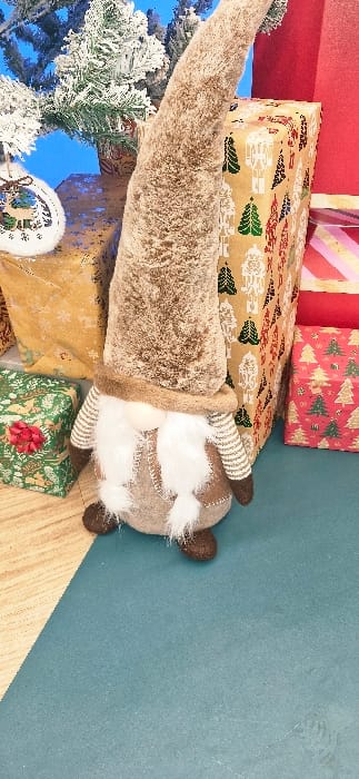 The scene depicts an indoor festive display located in Vendin-le-Vieil, France. A plush gnome figure is positioned centrally, featuring a tall, pointed, shaggy brown hat that covers its eyes, a small light-colored nose, and a prominent white beard. Its body is constructed from light brown fabric with dark stitching details, and it has striped arms with dark brown hands and rounded dark brown feet. The gnome stands on a teal-colored surface, which transitions to a light wood-grain floor on the left side of the frame. Surrounding the gnome are several wrapped gift boxes: a large vertical box wrapped in brown paper with white nutcracker and green Christmas tree patterns is to its right, alongside a smaller red box with green tree patterns. To the gnome's left, a gold-colored box with snowflake patterns is visible, and a green box with a small red bow rests further left. In the upper left corner, an artificial Christmas tree with snow-flocked branches and a white ornamental cutout of a deer hangs against a plain blue background. All subjects are inanimate and arranged in a static configuration. No human figures are present.