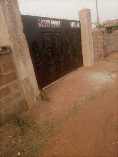 A brown metal gate, adorned with circular and spear-shaped ornaments, stands between two cement pillars. The left pillar is partially integrated with a brick wall. A brick fence runs along the right side of the gate. The ground in front of the gate is dirt, sparsely covered with small patches of grass and some dry leaves. A power line and a hint of a building are visible in the background, adding depth to the scene.