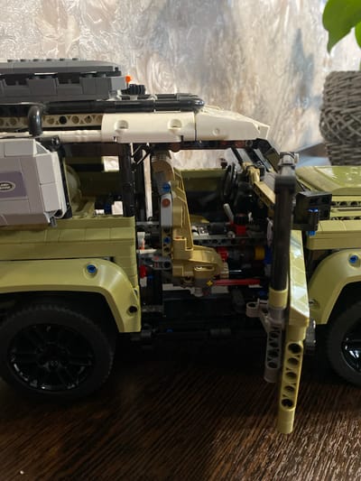 A detailed, forensic, matter-of-fact description of the scene:
The image displays a close-up, side view of a detailed LEGO Technic Land Rover Defender model, predominantly in a light green color. The model is situated on a wooden surface, likely a floor or table, with a textured, light-colored wall as the background. A wicker basket and a green plant are visible in the upper right portion of the background.
The vehicle model exhibits intricate mechanical details. The driver's side door is open, revealing the interior, which includes a tan-colored seat, a steering wheel, and various gears and mechanical components. The exterior features a roof rack with dark gray and black elements, a black snorkel on the driver's side, and a large black wheel with a silver rim. The rear section of the vehicle is partially visible, showing more light green panels and mechanical linkages. A white storage box with a purple "LAND ROVER" logo is attached to the side of the vehicle near the front.
There are no people present in the image. The scene appears to be an indoor setting. The lighting suggests it is daytime. The camera angle is at eye level with the vehicle. There is no indication of the specific location within the city of Берёзовая Роща, Russia, as the image contains no context clues to that effect.