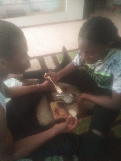 Two individuals, likely young adults with dark hair and skin, are engaged in sharing a meal in what appears to be a casual indoor or semi-outdoor setting in Oke Ogun, Nigeria. The scene is lit by diffused daylight.
The person on the left is wearing a light-colored top and a silver-toned bracelet on their left wrist. They are holding a gold-colored fork, ready to eat. The person on the right is wearing a patterned t-shirt, primarily white and grey, featuring a prominent green graphic or stylized text on the chest. While difficult to decipher precisely due to blurriness, it appears to contain letters, possibly resembling "TREV" or "TRVE".
Several small dishes and bowls are arranged on a surface in front of them, indicating a shared meal. One dish contains a fried item, possibly plantain or a fritter, while another small bowl holds a light-colored sauce or dip. A spoon is also visible among the cutlery. A mobile phone, encased in what appears to be a yellow-gold cover, is placed directly on the dining surface next to the food.
The background is softly blurred, showing lighter-toned walls or surfaces and hints of greenery, suggesting a view towards an outdoor area beyond a dark, slatted structure, possibly a fence or railing. The overall atmosphere suggests a relaxed, informal mealtime. No other discernible text is visible in the image.