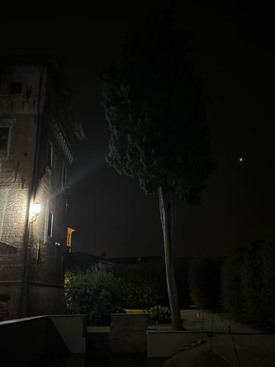 The image depicts a dark, nighttime scene featuring a tall, slender tree in the foreground, its silhouette partially illuminated by an unseen light source. To the left, a portion of a brick building with architectural details and an exterior light fixture is visible. The building appears old, with visible mortar and brickwork. Beyond the tree and building, dense foliage forms a dark, almost impenetrable background, suggesting a garden or natural area. The sky is dark and overcast, with a faint green light visible in the upper right quadrant, possibly indicating artificial lighting or an atmospheric phenomenon. The overall impression is one of stillness and nighttime ambiance. The location, within the city of Lagnasco, Italy, implies a European architectural style and potentially a historical setting. No people or animals are present in the frame.