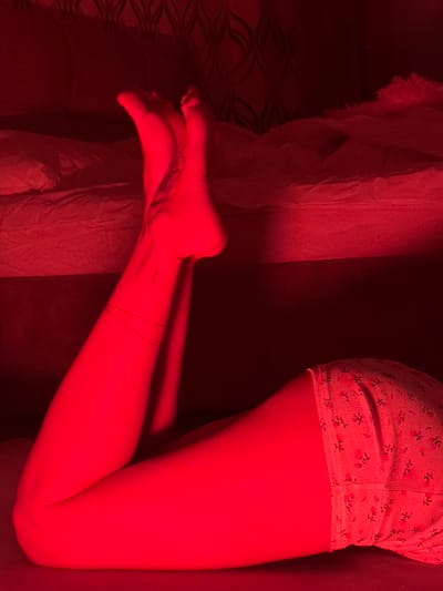 A close-up photograph taken in red lighting shows a person lying on a patterned surface, possibly a bed. The person is wearing floral-printed shorts. Visible body parts include the right leg, ankle, and foot. The foot has a tattoo on the instep, and the ankle has a thin object around it. A crumpled white fabric, likely bedding, is located to the left. The city of Svkh Voronezhskiy, Russia, is irrelevant to the visual content of the image.
