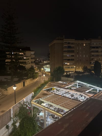 The image depicts a night view of an outdoor restaurant or patio area in Fuengirola, Spain. The foreground shows multiple levels of tables and chairs set under illuminated canopies. The middle ground features a street with a lamppost and a tall pine tree casting a shadow. Across the street, a multi-story building with lit windows and a sign reading "BLANCA SERVICE CENTER" is visible. Further in the background, other buildings and streetlights create a city skyline under a dark night sky. The overall impression is a typical urban night scene in a Mediterranean coastal town.