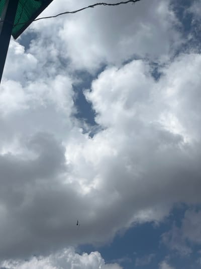 The scene, captured in Gitothua ward, Kenya, presents an upward-angled view of a daytime sky predominantly filled with dense, variably illuminated cumulus clouds, ranging from bright white to dark grey. Sections of clear blue sky are visible through gaps in the cloud cover. In the upper-left corner, a dark structural post and a fragment of a green corrugated roof are present. A dark, twisted cable traverses the upper portion of the frame from left to right. A small, dark-colored helicopter is observed in flight in the lower-middle left, partially enveloped by clouds. A narrow, light-toned object hangs vertically below the helicopter. No human figures are present.