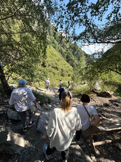 The image depicts a group of people hiking on a rocky, wooded trail in a mountainous region. The environment is characterized by lush green foliage, large rocks, and a steep hillside in the background. Sunlight filters through the trees, casting shadows on the path.

In the foreground, three individuals are visible from behind. The person on the far left wears a white t-shirt with a floral design and a light blue headscarf. They are carrying a light-colored bag over their shoulder. Next to them, to the right, is another person wearing a light-colored sweatshirt and dark pants, with blonde hair tied in a ponytail. To the right of this person is an individual in a white t-shirt and dark pants, with short dark hair.

Further up the trail, several other people are walking in the same direction. One person in a white t-shirt and striped pants is visible. Other individuals are scattered along the path, some further in the distance.

The overall scene suggests an outdoor recreational activity, likely a hike or trek, in a natural, undeveloped area. The location in the city of Chemal, Russia, is not directly discernible from the visual information alone, but the mountainous terrain and vegetation are consistent with areas found in Russia.