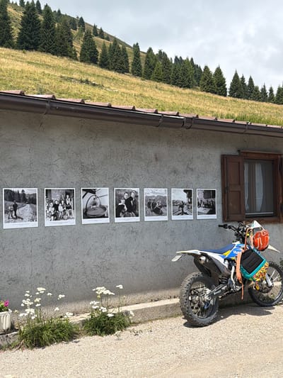 This is an eye-level, medium shot of a stucco building with a series of black and white photographs displayed on its exterior wall. The photographs appear to be historical in nature, depicting people in various rural settings. To the right of the photographs, a wooden-shuttered window is visible. In front of the building, a dirt bike is parked, equipped with an orange helmet and a colorful bag attached to its rear. The ground in front of the building is paved with gravel. Behind the building, a grassy hillside covered with trees rises steeply, under an overcast sky. White wildflowers are growing in front of the building. The location is identified as Lateis / Latais, Italy.