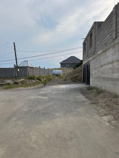 The image depicts an outdoor scene in Guliston, Tajikistan, featuring an unpaved road leading into a developing area. On the right side of the road, a partially constructed building made of grey brick and concrete is visible. Its walls are partially finished, and windows are open openings. To the left of the road, a brick fence encloses an area with a grey metal-roofed structure. Further in the background, another residential building with a dark pitched roof is situated on a hillside. A large, cylindrical metal tank, likely for water or fuel storage, sits on the ground near the fence. Overhead, power lines are strung between wooden utility poles. The ground is a mixture of dirt, gravel, and some patchy vegetation. The sky is overcast with scattered clouds. The overall impression is one of ongoing construction and development in a semi-urban or rural setting.