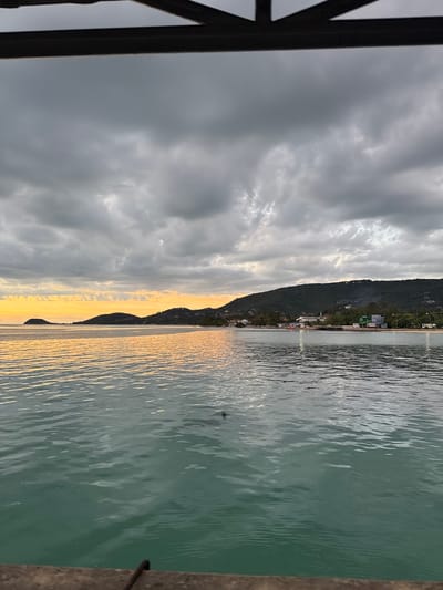 This media file captures a view of the sea from a pier or dock in Nathon, Thailand. The image is oriented vertically. On the left side of the frame, a dark, weathered wooden structure, likely part of the pier, occupies a significant portion. Beyond this structure, the sky is filled with dramatic, dark gray clouds, suggesting overcast or stormy weather. Streaks of sunlight break through the clouds, creating a contrast of light and shadow.
The scene opens up to a calm sea. The water is a muted green color, reflecting the overcast sky with subtle ripples on its surface. Along the horizon, a dark, tree-covered mountain range forms the backdrop for a small coastal town. Buildings are scattered along the base of the mountain, some appearing as lights, hinting at activity even in the subdued light. The water near the shore is illuminated by a warm, golden glow, indicating that the sun may be setting or rising, casting a soft light.
There are no people visible in the image. The focus is on the natural landscape and the man-made pier. The overall mood is serene, with a touch of dramatic flair from the cloudy sky. The location cues, such as the palm trees implied by the lush mountain vegetation and the style of the buildings, along with the provided location context, point to a tropical coastal setting. The time of day is likely dawn or dusk, given the warm light on the water. No discernible text is visible within the image.