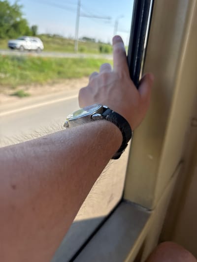 The media file shows a close-up of a person's arm and hand, viewed from a slightly high angle, as if looking out of a vehicle window. The person is wearing a black watch with a silver-toned case on their left wrist. Their hand is resting on the edge of what appears to be a window frame, with fingers extending slightly over the glass. The background, visible through the window, is blurred but suggests an outdoor scene with green foliage and a road. A white car is visible in the distance. The lighting indicates it is daytime. The scene appears to be a still moment, possibly during travel. There is no visible text.