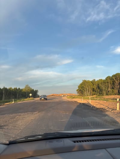 The image depicts a roadside construction scene in Plodopitomnik, Russia. A black car is visible on an asphalt road that appears to be under construction, with piles of dirt and gravel on the side. A line of trees, primarily deciduous with some evergreens, borders the construction area. The sky is a clear blue with wispy white clouds, suggesting it is daytime, likely late afternoon given the long shadows. Several road markers, white with red tops, are placed along the roadside. The perspective suggests the photo was taken from inside a vehicle, possibly looking out of a side window, with part of the vehicle's interior visible in the foreground. The overall scene conveys a sense of ongoing development and transition in a rural or semi-rural setting. No text is visible in the image.