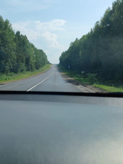 The image shows a view from inside a vehicle looking out at a two-lane asphalt road curving gently uphill. Dense green trees line both sides of the road, creating a tunnel effect. The sky is partly cloudy with bright sunlight. A blue and white parking sign is visible on the right side of the road, near a patch of overgrown vegetation. The foreground is dominated by the dashboard or interior of the vehicle, which is out of focus and appears to be a dark gray material. The scene suggests a drive through a wooded area, possibly in the countryside or a rural part of Morovo, Russia, given the context. There are no people visible in the image.