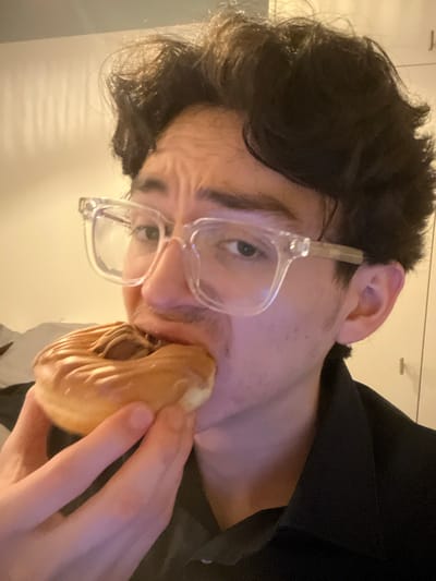 A man with dark, curly hair and clear glasses is holding a chocolate-filled pastry to his mouth, seemingly about to take a bite. He's wearing a black shirt or jacket. The photo is taken at a slightly skewed angle, and the background appears to be a simple, light-colored wall. The focus is mainly on the man's face and the pastry.