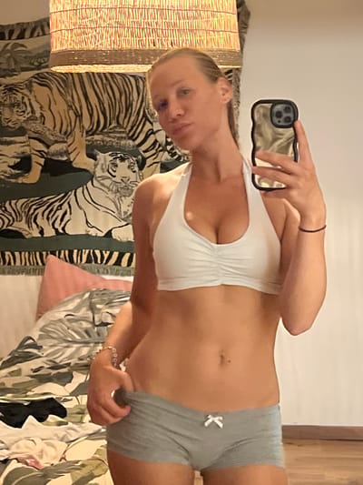 The image shows a person taking a mirror selfie in what appears to be a bedroom. The person is wearing a white sports bra and grey shorts. They are holding a smartphone in their right hand, which is reflecting the image. The background includes a woven lamp, a tapestry with a tiger print, and some foliage. The lighting suggests it is daytime. The person's expression is neutral.