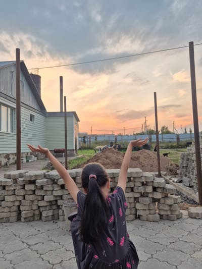 A child with long dark hair tied in a ponytail, wearing a dark dress with pink butterfly patterns, stands in a construction area with their arms outstretched, facing away from the camera. In the foreground, stacks of grey concrete paving stones are arranged, and the ground is paved with similar stones. Behind the child, a light green building with a white roof is visible, along with construction materials, mounds of dirt, and vertical metal poles. In the background, a fence with power lines and utility poles stretches across the horizon. The sky is a blend of grey clouds and a warm orange and pink sunset. The overall setting appears to be the outskirts of Новоишимское, Kazakhstan, possibly a residential construction site.