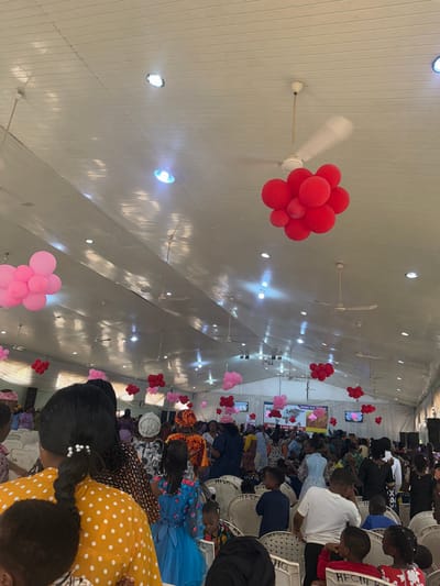 The image shows an indoor event, likely in a hall or large room in Barnawa, Nigeria, indicated by the presence of a sign visible in the background. Numerous people, including women and children, are seated on white plastic chairs. Red and pink balloons are scattered throughout the room, suspended from the ceiling. The ceiling is white and features multiple recessed lights and ceiling fans. Many people are looking down.