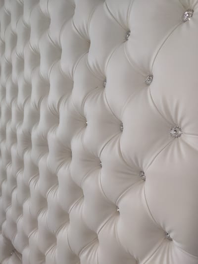 The image shows a close-up of a tufted white upholstery, likely a headboard or wall paneling, with diamond-like accents. The material appears to be faux leather and is arranged in a diamond pattern with deep button tufting, creating a plush and luxurious texture. The lighting highlights the curves and folds of the fabric, with the diamond accents catching the light and adding a subtle sparkle. The perspective is from a low angle, looking upwards at the surface. The scene does not contain any people or specific environmental cues that would indicate a location within Supsekh, Russia.