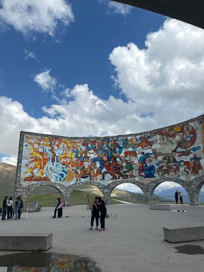 The image depicts a large, curved mosaic mural on a concrete wall, depicting numerous figures engaged in various activities, possibly representing historical or mythical scenes. The mural is located outdoors in Gudauri, Georgia, as indicated by the mountain landscape visible through arched openings beneath the mural. The sky above is blue with scattered white clouds.
In the foreground, a paved plaza is visible, with several people scattered throughout. On the left, a group of five individuals stands near a white structure and a railing. In the center, a person is seated on a black object, facing the mural. Further to the right, two individuals stand close together, with one appearing to take a photo or video. Another person stands further to the left in the foreground.
The environment suggests a scenic overlook or monument. The ground is paved with rectangular stones, and there are concrete benches and some puddles on the ground, reflecting parts of the mural. The background features rolling hills and distant snow-capped mountains, confirming the mountainous location. The overall impression is a public space designed to showcase art and offer panoramic views.
