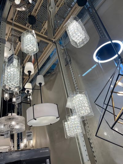 The image displays an indoor retail setting, specifically a section dedicated to lighting fixtures, located in Midwest City, United States. The scene is dominated by numerous hanging chandeliers and pendant lights suspended at various heights.
Key objects include:
- Multiple crystal-style chandeliers, characterized by clear cylindrical or rectangular outer glass or acrylic shells containing an array of small, reflective, faceted crystal-like elements. Several of these feature yellow price tags, with visible prices including "$199.00", "$149.00", and "$139.00".
- A large, prominent drum-style chandelier with a wide, circular white fabric shade framed by black metal supports, hanging centrally in the lower-middle portion of the image.
- A black-framed chandelier with multiple arms extending horizontally, each terminating in a white, inverted conical glass shade.
- Geometric light fixtures, including a large, open-frame structure composed of black metal in the shape of a cube or pyramid, located on the right side.
- Other varied light fixtures, including smaller glass and metal designs, are visible in the background and foreground.
The environment features a high ceiling with visible metal grating and industrial-style metal shelving or racking structures in the background, suggesting a warehouse or large retail store environment. The walls appear to be a neutral light gray. A black product box labeled "CHANDELIER" and bearing the number "1012214988" is partially visible in the lower left corner. No individuals are present in the image.