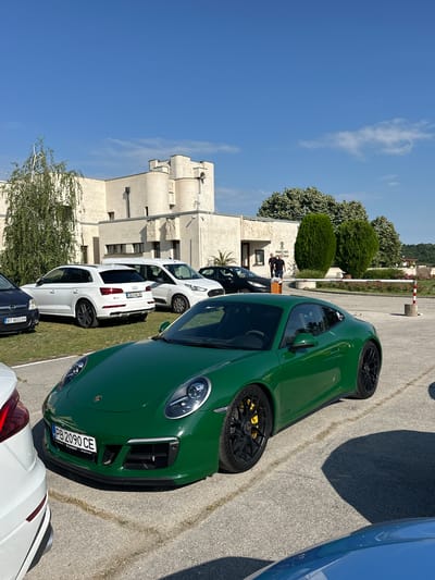 A front view of a green Porsche 911 GTS is parked in a paved lot in front of a beige, castle-like building under a clear blue sky. The car has yellow brake calipers and black wheels. To the left of the Porsche is a white Audi Q5 SUV. In the background, a white van and a black car are also parked. Two men are standing near the entrance of the building, which has decorative carvings. The location is the Church of the Nativity of Christ in Arbanassi, Bulgaria.