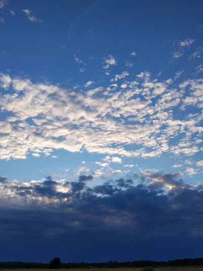 A vertical, eye-level shot showcases a dramatic skyscape over Ustyvytsya, Ukraine, at dusk. The sky is a deep blue, transitioning to lighter hues near the horizon. A band of bright white cumulus clouds stretches across the middle of the frame, interspersed with smaller, wispy clouds. Below this, a darker, heavier layer of clouds obscures the setting sun, with patches of light breaking through. The bottom of the image reveals a dark silhouette of a treeline and a flat, indistinct landscape, suggesting a rural or open area. No people or specific structures are clearly identifiable.