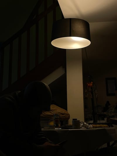 The image portrays a dimly lit indoor scene in Villeurbanne, France. A lone individual, silhouetted and clad in a dark hooded garment, is seated in the foreground with their head bowed, holding a small electronic device. A light-colored tablecloth covers a table directly in front of the person, displaying a glass containing liquid, a white box, and a mug, alongside other unidentifiable items. A black drum-shade floor lamp stands prominently to the right of the individual, providing localized illumination downwards. In the background to the left, a wooden staircase with dark balusters ascends, mostly obscured by shadow. A cream-colored vertical support structure or wall is visible beneath the lamp. Further right in the background, a dark-framed object hangs on a wall, with a decorative item featuring multiple dangling elements attached to a vertical stand near the lamp.