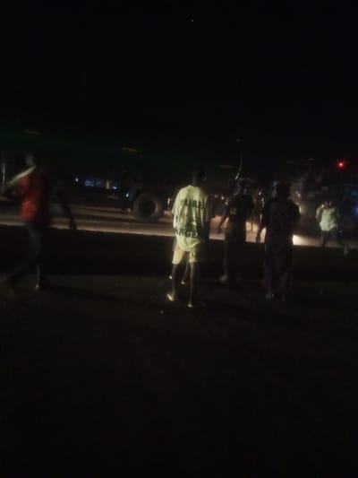 This is a nighttime, low-light, and blurry photograph taken on a road in Nassarawa, Nigeria. Several people are visible, mostly blurred and indistinct due to the low light and motion. In the foreground, a person in a white t-shirt with text and light-colored shorts is standing. To the left, another person in a red shirt is also visible. In the background, a large truck with its headlights on is partially visible, along with other figures and lights that suggest activity or a gathering. The scene appears to be a roadside, possibly in a semi-urban or rural area, given the presence of the truck and the unpaved or rough surface. The overall impression is one of a spontaneous roadside stop or encounter at night.