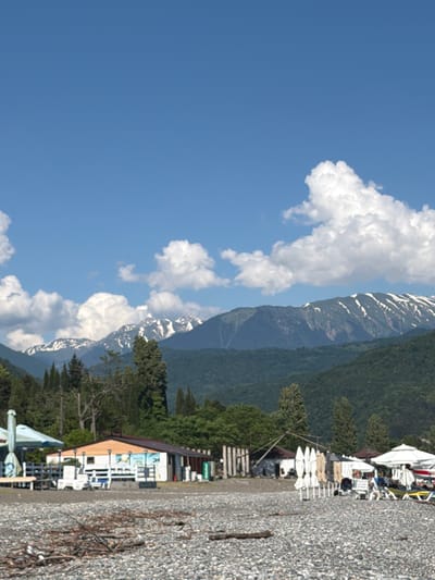 The image depicts a view of the coastline in Amzara, Abkhazia. In the foreground, a pebbled beach is visible with scattered driftwood. Further back, several beach umbrellas and lounge chairs are set up, suggesting a recreational area. A low-rise building, possibly a cafe or a shop, with an orange-tiled roof and white walls adorned with murals, is situated behind the beach furniture. The midground features dense green trees, and the background is dominated by a majestic mountain range with snow-capped peaks under a clear blue sky dotted with fluffy white clouds. The scene conveys a peaceful, natural landscape combined with amenities for beachgoers.