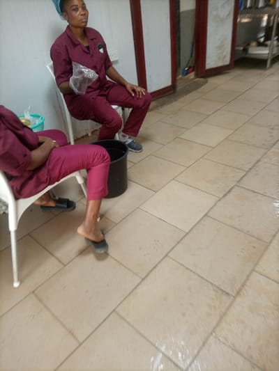 This image depicts an indoor scene in the city of Opete, Nigeria, featuring two individuals seated in what appears to be a break area or utility room. The floor is covered with light-colored square tiles, many of which exhibit wet patches and streaks. The left wall is white, while the background features two reddish-brown wooden door frames. The rightmost door is partially open, revealing a darker interior with a blue mop and a multi-tiered metal shelving unit.
The two individuals are adult females, both wearing maroon short-sleeved scrub-like uniforms with matching trousers.
The individual on the left, further into the background, has dark skin, with her hair covered by a light blue hairnet. She is seated on a white plastic chair and holds a clear plastic bag in her right hand. She is wearing grey and white athletic-style shoes. A wall power outlet is visible on the white wall behind her.
The individual in the foreground, partially cropped, also has dark skin and is seated on a white plastic chair. A black bucket is positioned on the floor between her and the other individual. A light blue plastic basket containing a clear plastic bag with light-colored contents is visible near her left foot. Her left foot is inside a black slide sandal, while her right foot is partially out of another black slide sandal, with the sole of her foot making contact with the sandal.