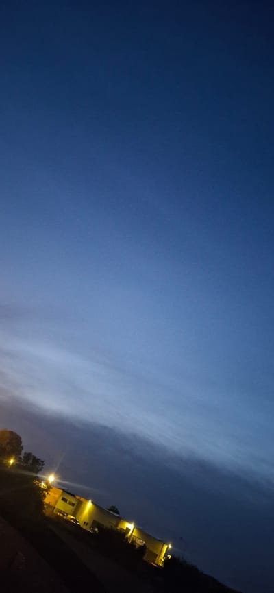 A diagonal shot of an industrial-style building at dusk or night in Sant'Egidio alla Vibrata, Italy. The sky is a deep, clear blue that fades to a lighter shade near the horizon, with faint wispy clouds visible. The building, illuminated by bright yellow lights, appears to have a light-colored facade and a dark roofline. Several windows are visible, some with lights on. Streetlights are also present, casting pools of light. Trees and vegetation frame the building. The overall impression is one of a quiet, commercial area as day turns to night.