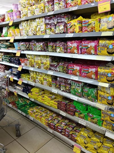 The image captures a brightly lit aisle in what appears to be a supermarket or grocery store, focusing on shelves heavily stocked with instant noodle packages. The scene is dominated by multiple rows of shelving extending into the background, all filled with various brands and flavors of instant noodles.
A metal shopping cart is partially visible on the left side of the frame, its handle and wheels prominent, suggesting a customer's perspective or an active shopping environment. The floor is tiled with light-colored, possibly beige or white, square tiles.
The instant noodles are neatly arranged on multiple grey shelves. Prominent brands include "Indomie" and "Nala".
"Indomie" packages are seen in several variations:
* Red and pink packages often depict cooked noodles with fried chicken or prawns and an egg, frequently labeled "Instant Noodles" and "Mi Goreng". Some have "5" indicating a pack quantity.
* Brown or gold packages often show beef, with "Beef" visible on some. Other brown packages show "Kari Ayam" (chicken curry).
* Yellow packages are also present, often featuring chicken imagery.
* Green packages are visible in some sections, indicating other flavors.
"Nala" brand packages are predominantly yellow, featuring a bowl of noodles and often the text "Instant Noodles", "Chicken Flavour", "Value Pack", and "FREE TTE INSIDE" (likely "FREE TASTE" or "FREE TRAY"). These also frequently display "5" for pack quantity.
Various price tags, typically yellow or orange, are attached to the edges of the shelves. One tag near the top right shows "99/-" and "21 February 2020", suggesting a specific date for a sale or price validity. Other smaller white labels with black text are also attached to the shelves, likely containing product codes or prices.
The overall impression is one of a well-organized and well-stocked instant noodle section, ready for shoppers. No people are visible in the image, allowing the focus to remain solely on the product display.