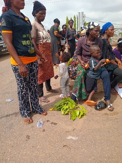 The scene is a public gathering of mostly women and children in Ungwan Tanko, Nigeria, at the intersection of Latitude: 10.422 and Longitude: 7.49. It is a dusty, paved street with scattered debris. A woman wearing a black shirt with the words "In This World" and black and white pants with the word "love" is standing near the center of the image. She is facing the camera. Other women in various clothing and headscarves are standing and sitting. There is a group of children, including a young boy wearing a blue shirt. They are sitting on the ground or standing, interacting with each other and the adults. There is a large pile of green leaves lying on the ground. The overall impression is one of a bustling public space with a diverse group of people going about their day.