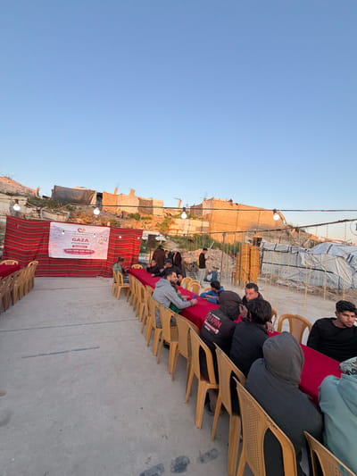The image depicts an outdoor gathering set on a concrete ground surface in Jabalia, Palestinian Territory, under a clear, light blue sky. In the foreground, multiple long, narrow tables draped with red tablecloths are arranged parallel to each other. Numerous light brown plastic chairs are positioned around these tables. Approximately two dozen men are visible, predominantly seated at the tables, with some standing in the background.
To the left, a white banner with a red border and heart logo is prominently displayed against a red and white striped fabric. The banner reads "HELP A NEEDY," "GAZA," "#IFTAR MEALS," and includes a phone number: "Info: 939-636-0672." String lights with exposed white bulbs are suspended overhead, illuminating the area. The background reveals a landscape of severe structural damage and makeshift constructions. Rubble, damaged concrete structures, exposed rebar, and temporary shelters made from tarpaulins and various materials are visible on an elevated, uneven terrain. Additional temporary structures or stacked materials are present on the right side of the frame. The men at the tables appear to be waiting or conversing, while a few individuals in the mid-ground are standing near the damaged structures.