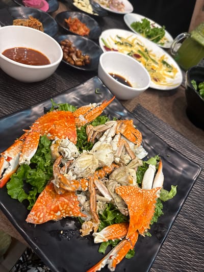 The image depicts a table spread with various dishes, primarily focusing on a large black square plate filled with what appears to be cooked crab meat arranged on a bed of lettuce. The crab shell is a speckled orange color. Other dishes on the table include small bowls containing sauces and condiments. There is also a partially eaten dish with green and yellow vegetables. A glass of green juice is also visible. The table has a textured, dark-colored surface. The photograph appears to have been taken indoors. The image does not provide information about a specific location in Kathmandu Metropolitan City, Nepal, nor does it show people, actions, or interactions.