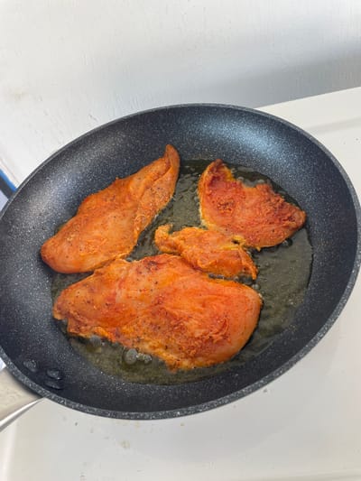 The image shows three pieces of seasoned chicken breast being pan-fried in a dark gray frying pan. The chicken is coated in a reddish-orange seasoning blend, likely paprika-based, with visible specks of black pepper and herbs. The chicken is cooking in oil, which is bubbling around the pieces.
The frying pan is dark gray with a textured, non-stick surface. The pan has a metallic handle. The background is a bright white surface, possibly a countertop or stovetop, with a faint texture. The lighting suggests an indoor setting, likely a kitchen. There are no visible people or animals. The scene depicts the process of cooking food.
There is no visible text in the image. The time of day and weather are not discernible. The location context of Skala Avdiron, Greece, is provided but not visually evident in the image itself. The overall impression is one of meal preparation.