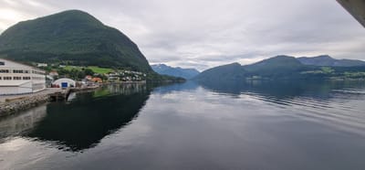 The image depicts a serene waterscape with a village nestled against lush green mountains, likely in Volda, Norway. In the foreground, a calm body of water reflects the overcast sky and the surrounding landscape. On the left side of the image, a prominent building complex with a white facade and numerous windows is situated along the waterfront. Adjacent to this building is a dock with a white arched structure, and a few small vehicles are visible. The buildings of the village, characterized by colorful roofs, are scattered along the base of a steep, tree-covered mountain. Further in the distance, other mountains rise, some with snow-capped peaks, under a cloudy sky. The overall scene conveys a sense of tranquility and natural beauty.