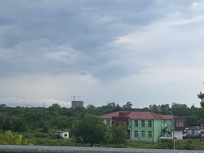 The image depicts a cloudy sky over a rural landscape with buildings and dense vegetation. In the foreground, a horizontal metallic railing is visible, suggesting the viewpoint is from a bridge or elevated structure. Behind the railing, lush green trees and bushes dominate the middle ground.
Two distinct buildings are present. The larger building, painted in a light green with a red roof, features multiple windows and appears to be multi-storied. Adjacent to it, a smaller building with a maroon roof is partially visible, connected to the green structure. Further to the left, a small white structure with a red roof stands amidst the foliage.
In the background, a cluster of trees forms a dense canopy. Beyond the trees, a construction site with a tall building and a crane is visible, indicating ongoing development. In the distance, a cloudy sky with diffused light covers the scene. The overall impression is of a semi-urban or suburban environment with elements of both nature and construction, located in Nakaidzeebi, Georgia. No people are visible in the frame.
