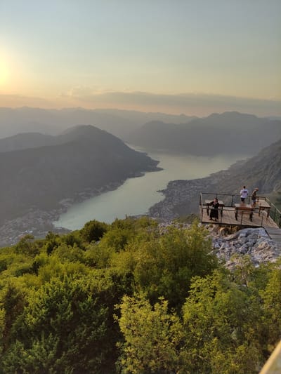 A high-angle shot captures a breathtaking panorama of a river valley and the surrounding mountains at sunset. The scene is dominated by a lush green forest in the foreground, with a wooden observation deck jutting out over the cliff edge. On the deck, three individuals are visible, enjoying the view. One person stands near the railing, looking out at the expansive landscape, while two others are seated at a wooden table.
Below the observation deck, the river meanders through a narrow valley, with a small town nestled along its banks. The buildings of the town are densely packed, giving a sense of scale to the vastness of the natural surroundings. The mountains in the distance are layered in muted tones of gray and blue, softened by the haze of sunset. The sky transitions from a pale yellow near the horizon, where the sun is setting, to a soft blue overhead. The overall mood is one of tranquility and awe, enhanced by the warm, golden light of the setting sun. The location appears to be Žanjev Do, Montenegro, a place known for its dramatic natural beauty. There is no visible text in the image.