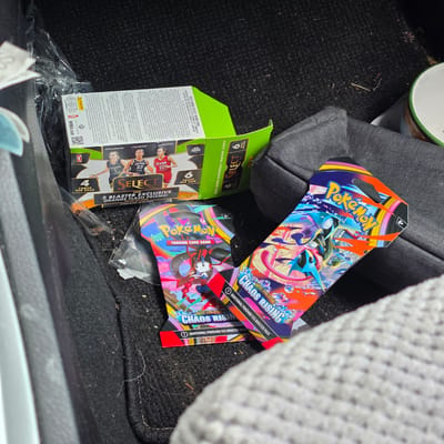 This media file shows two boxes of trading cards and two Pokémon booster packs resting on a dark, textured surface, likely the floor of a vehicle, with a glimpse of a gray seat in the background. The trading card boxes are for "2025 WNBA SELECT BASKETBALL" and are primarily green with images of basketball players like Caitlin Clark, A'ja Wilson, and Paige Bueckers. One box is open, revealing more details about the set's contents, including terms like "Silver Prizms," "Flash," and "Signature." The Pokémon booster packs are vibrant and feature artwork from the "Chaos Rising" set, with prominent Pokémon characters and the "Mega Evolution" branding. One booster pack is partially visible, showing "ADDITIONAL POKÉMON TCG BOOSTER PACK" text. The scene appears to be indoors, possibly in a car, given the textures and the presence of the seat. There is no indication of the time of day or specific weather. The overall impression is one of collectible items casually placed in a vehicle.