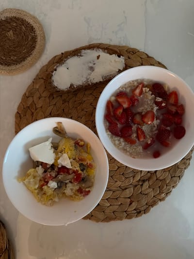 A close-up, overhead shot shows two white bowls of food and a slice of bread with cream cheese on a woven placemat. The bowl on the left contains scrambled eggs with red bell peppers, mushrooms, and feta cheese. The bowl on the right contains oatmeal topped with sliced strawberries. A dark bread slice with a generous spread of cream cheese sits on the placemat. The background is a white, textured surface, possibly a countertop, with hints of gray marble. A circular woven coaster is visible in the upper left corner. The scene depicts a breakfast spread. The location of Semiluki, Russia is not visible or inferable from the image.