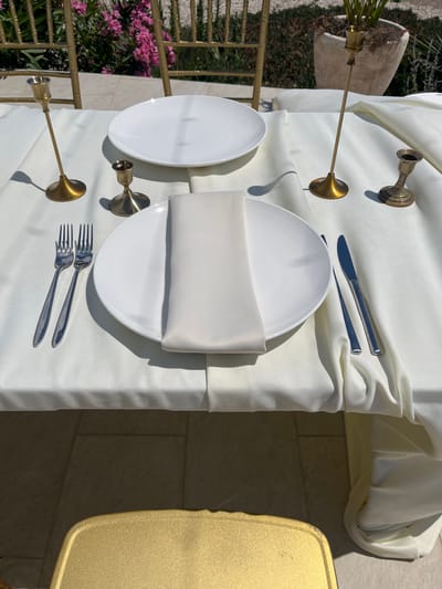 This is a top-down, eye-level medium shot of a formal dining table setting outdoors in Lastva Grbaljska, Montenegro. The table is covered with a white tablecloth. There are two place settings. Each setting includes a white plate with a folded white napkin on top, a fork to the left of the plate, and a knife to the right. Two brass candlesticks are positioned on the table, one behind each place setting. One candlestick is tall and slender, while the other is shorter and thicker. In the background, golden chairs are visible, along with pink flowers and greenery. The ground is paved with light-colored tiles. The lighting suggests it is daytime, with strong shadows cast by the candlesticks.