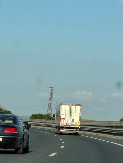 The image shows a sunny day on a highway. In the foreground, a black sedan is driving on the right lane of a multi-lane road. The sedan's brake lights are illuminated. In the middle ground, a white semi-trailer truck is traveling in the same lane as the sedan. The truck has its rear lights on. In the background, a pylon with power lines is visible, and beyond that, trees and hills can be seen. The sky is clear blue with a few white clouds. The highway appears to be in the vicinity of Yana, Bulgaria.