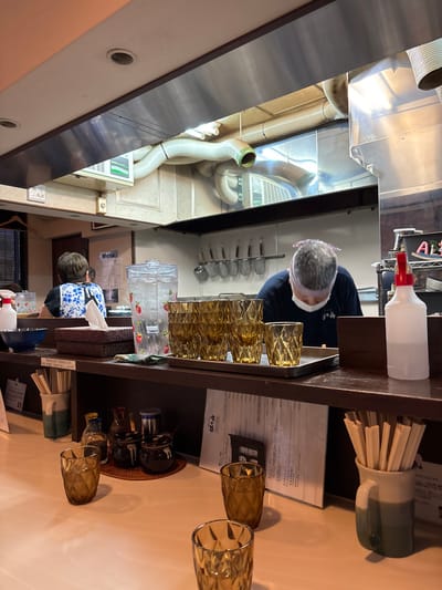 The image shows the interior of a small Japanese restaurant. It is located in Chūō, Japan at the coordinates (latitude: 35.687, longitude: 139.785). The scene is a kitchen counter with a customer sitting at the counter, and a chef preparing food. The chef is wearing a white headband and black shirt, and is using a large metal spatula to stir a pot on the stove. The customer is sitting at the counter with a cup of tea in front of them. There are several empty glasses on the counter, and several bottles of condiments in front of the customer.  The kitchen is equipped with a large stainless steel exhaust hood. The back wall is tiled in white, and the countertop is made of wood. The floor is tiled in black and white. The overall ambiance of the space is casual and homey.